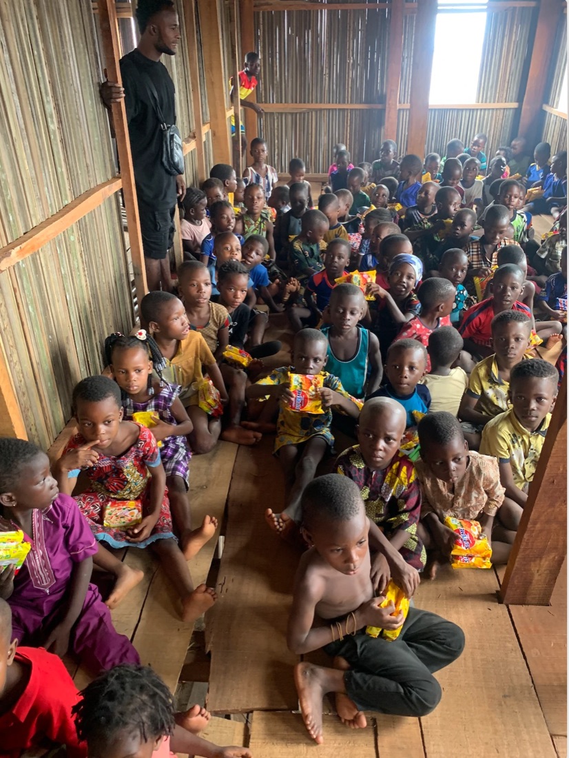 Children seated together on a wooden floor, many holding packaged snacks